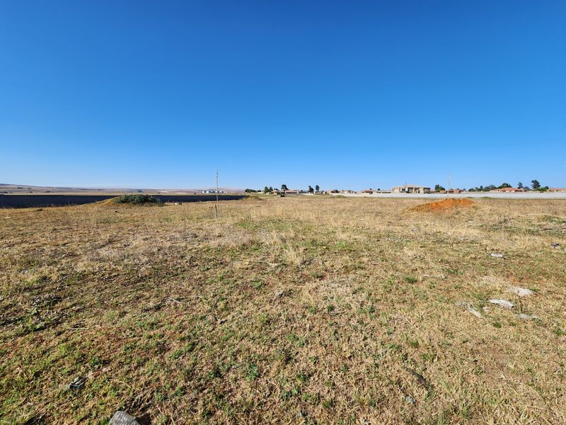 A wide-angle view of a vacant commercial land plot featuring dry grass and distant boundary fencing under a clear blue sky.