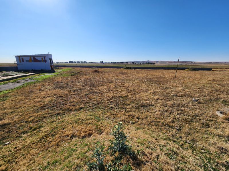 Expansive vacant commercial land featuring dry grass, a boundary wall, and a small existing structure on the property perimeter.