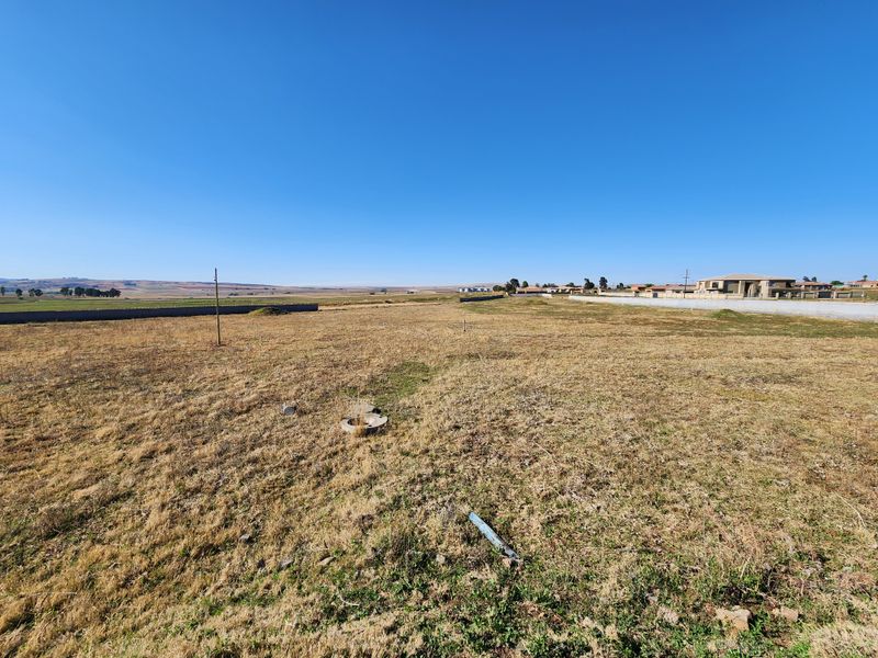 A wide-angle view of a vacant commercial land plot featuring dry grass, a perimeter wall, and clear horizon views.