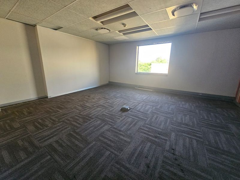 An empty office space featuring a suspended ceiling with recessed lighting and dark patterned carpet tiles with a central floor power outlet.
