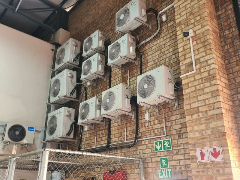 Industrial utility area featuring a wall-mounted array of air conditioning condenser units, exposed brickwork, and essential safety signage.