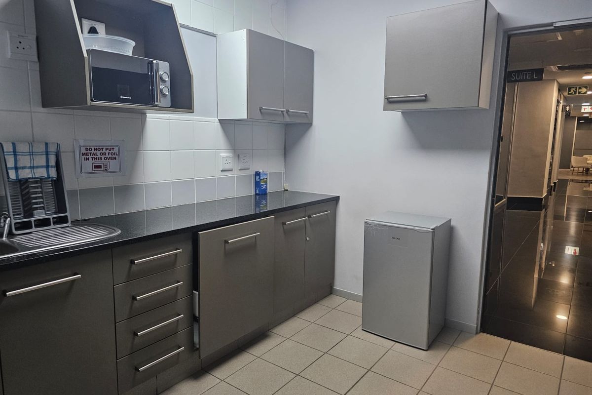 A functional office kitchenette featuring grey built-in cabinetry, a dark laminate countertop, stainless steel sink, and tiled backsplash.