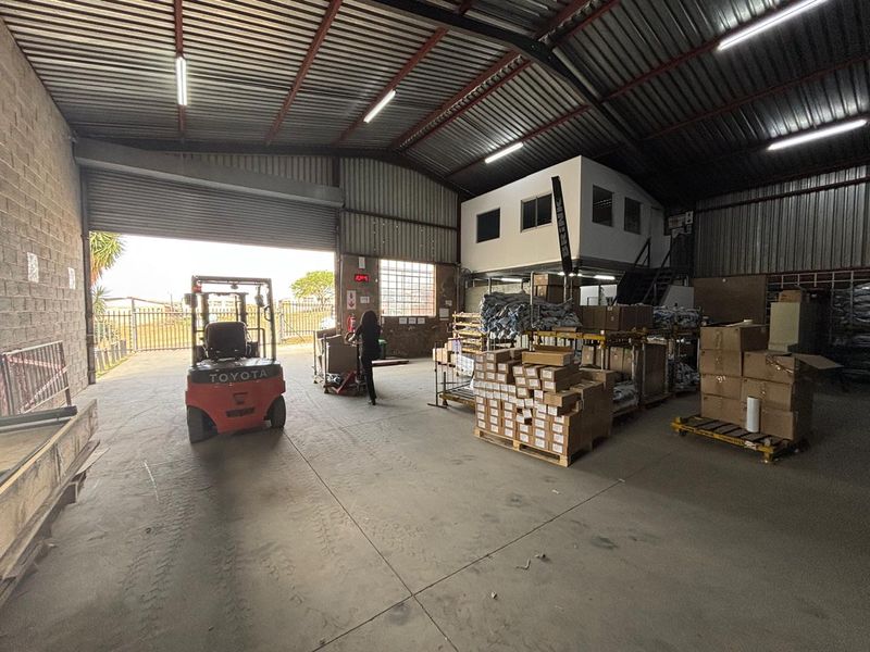 Spacious industrial warehouse featuring high ceilings with exposed steel trusses, concrete flooring, a large roller shutter door, and an integrated mezzanine office level.