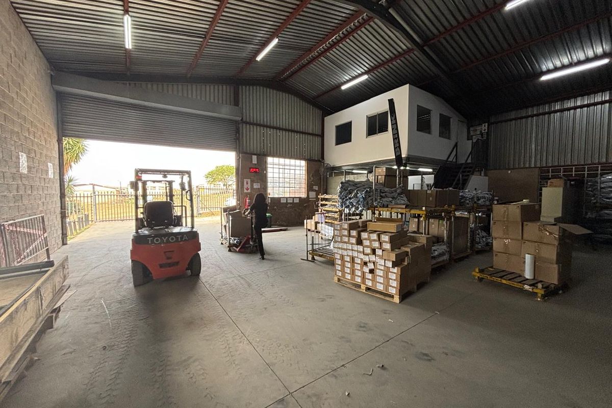 Spacious industrial warehouse featuring high ceilings with exposed steel trusses, concrete flooring, a large roller shutter door, and an integrated mezzanine office level.