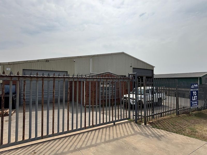 Industrial property featuring a warehouse with corrugated metal cladding, roller shutter doors, and secure steel palisade fencing on a concrete driveway.