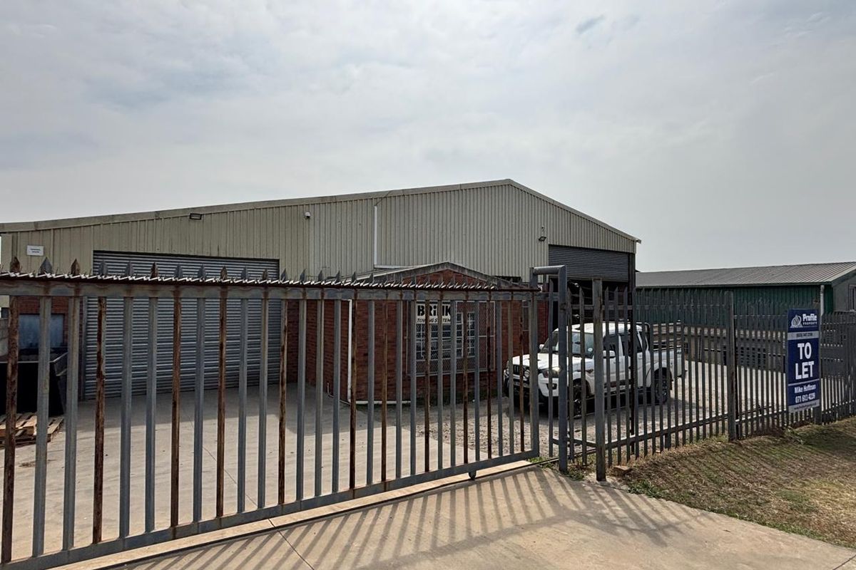 Industrial property featuring a warehouse with corrugated metal cladding, roller shutter doors, and secure steel palisade fencing on a concrete driveway.