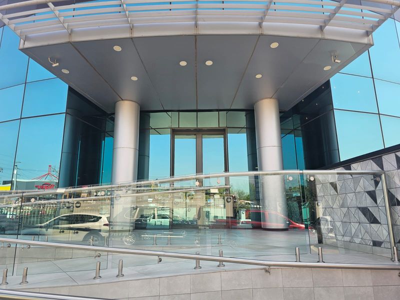 Modern office building entrance featuring a glass curtain wall facade, metal canopy, and a tiled accessibility ramp with a glass balustrade.