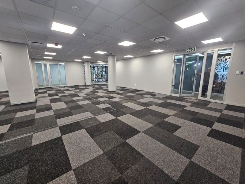 A spacious, open-plan office area featuring modern carpet tiles, a suspended ceiling with recessed lighting, and glass partition doors.