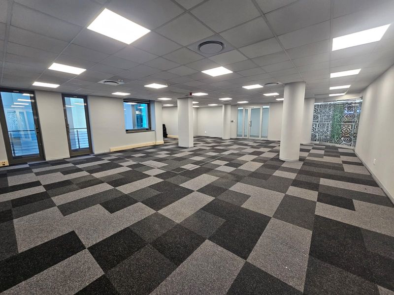 A spacious, open-plan office area featuring modern carpet tiles, recessed ceiling lighting, structural columns, and glass partition doors, offering a clean, professional workspace.