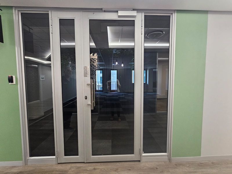 A modern office entrance featuring an aluminium-framed glass door with integrated access control, leading into a carpeted workspace with recessed ceiling lighting.