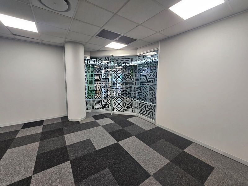 An empty office space featuring a modern glass partition with decorative film, carpet tile flooring, and a suspended ceiling with recessed lighting.