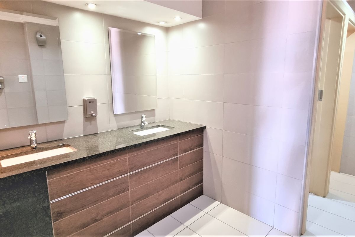 A clean, modern office restroom featuring a double vanity with granite countertops, wood-finish cabinetry, and tiled walls and flooring.