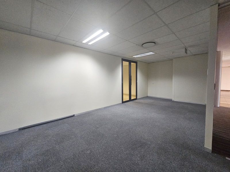 A vacant, open-plan office space featuring neutral walls, grey carpeting, and a suspended ceiling with integrated recessed lighting.