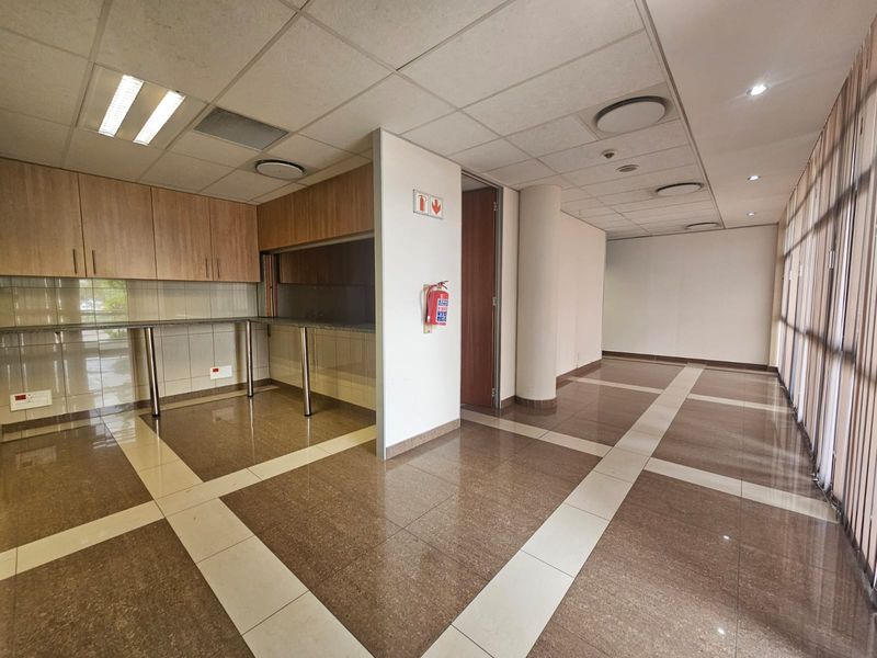 A spacious, vacant office area featuring a built-in kitchenette with granite countertops, tiled flooring, and a suspended ceiling with recessed lighting.