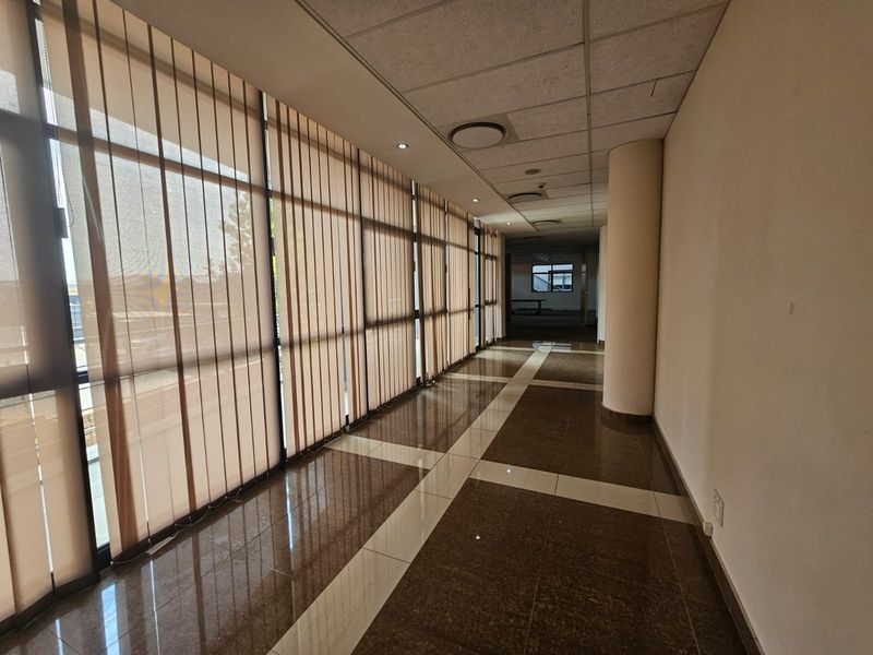 A spacious, well-lit office hallway featuring polished tiled flooring, a suspended ceiling, and large windows with vertical blinds.