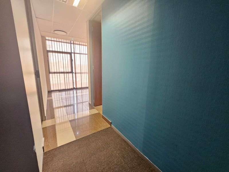 A clean, empty office hallway featuring a combination of tiled and carpeted flooring, a suspended ceiling, and a blue accent wall.