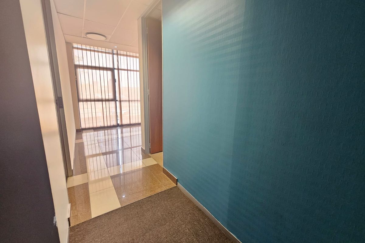 A clean, empty office hallway featuring a combination of tiled and carpeted flooring, a suspended ceiling, and a blue accent wall.