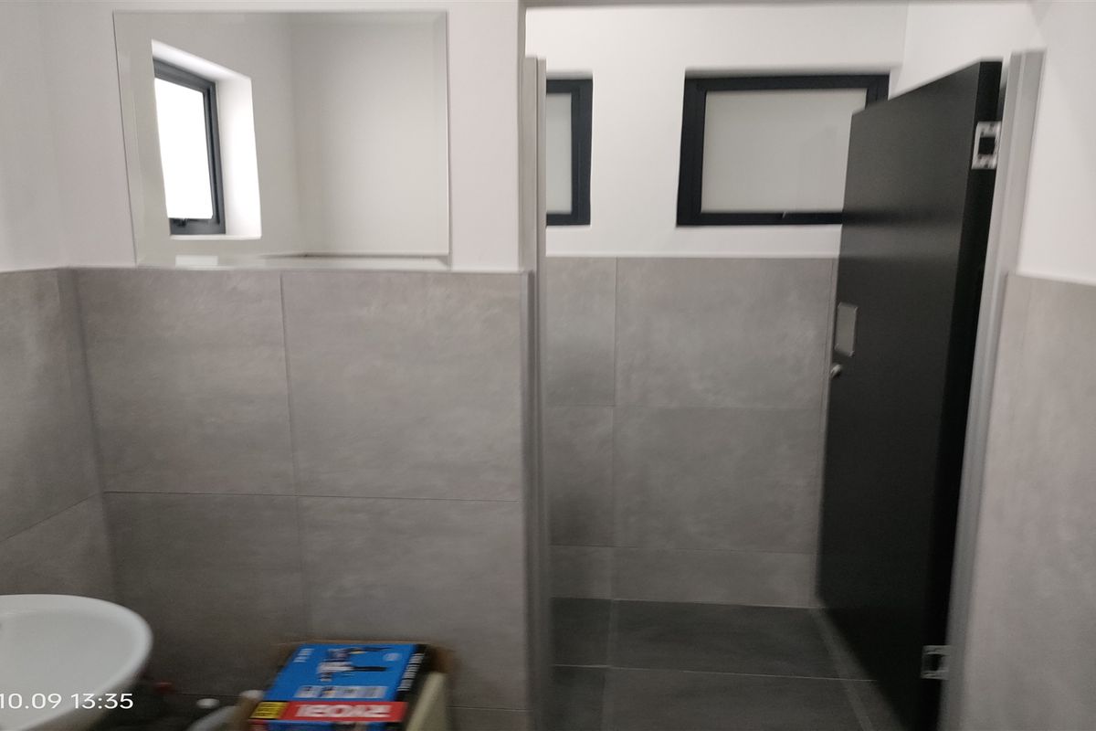 A modern industrial-style bathroom featuring grey wall and floor tiling, a wall-mounted basin, and a dark door.