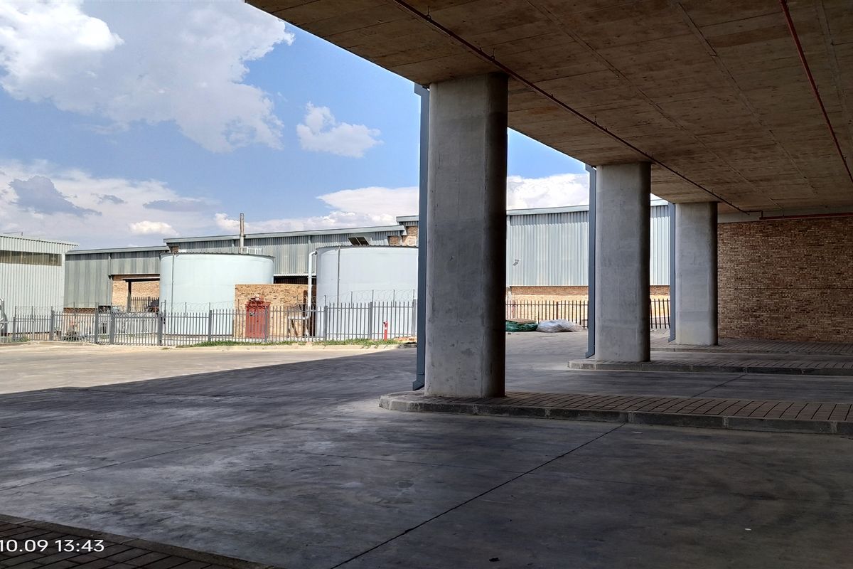 A covered industrial loading or parking area featuring concrete columns, a concrete ceiling, and a paved surface, with external storage tanks visible in the background.