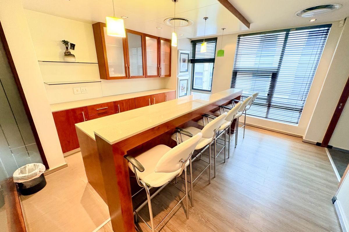 A modern office kitchenette featuring built-in wooden cabinetry, a stone-topped breakfast bar, and large windows with fitted blinds.