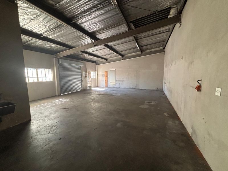 Spacious industrial utility area featuring concrete flooring, exposed steel roof structure, roller shutter door access, and basic wall-mounted electrical and plumbing fixtures.