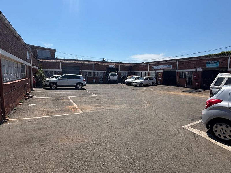 Industrial courtyard featuring an asphalt parking area, brick building facades, and multiple roller shutter access points for commercial or warehouse use.