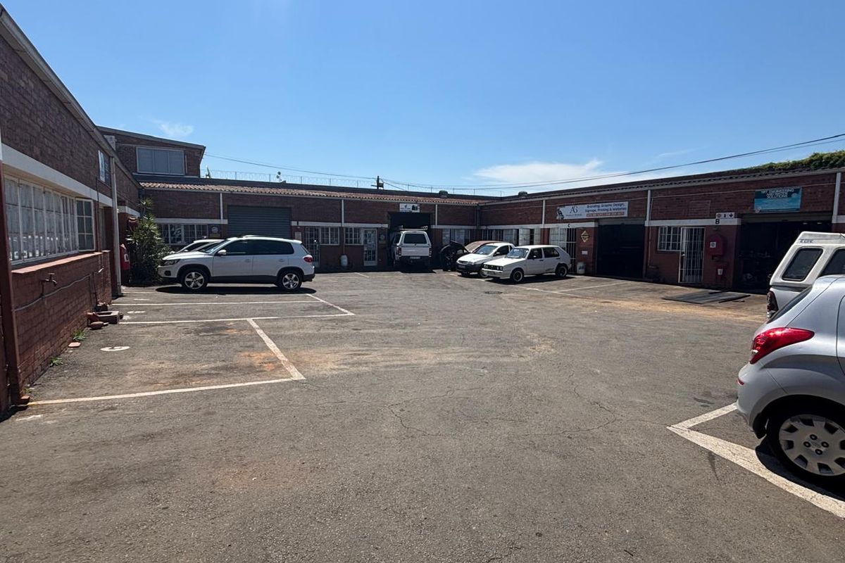 Industrial courtyard featuring an asphalt parking area, brick building facades, and multiple roller shutter access points for commercial or warehouse use.