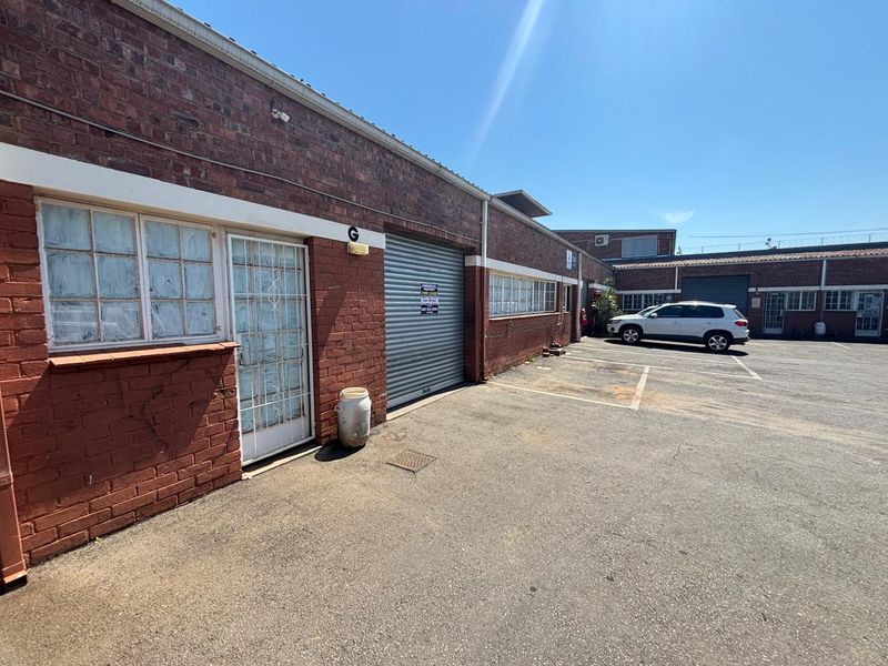 Industrial unit exterior featuring a roller shutter door, security gate, and brick facade with dedicated parking space in a paved yard area.