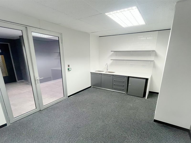 A clean, modern office space featuring a compact kitchenette with built-in cabinetry, sink, and floating shelves, finished with neutral tones and carpet flooring.