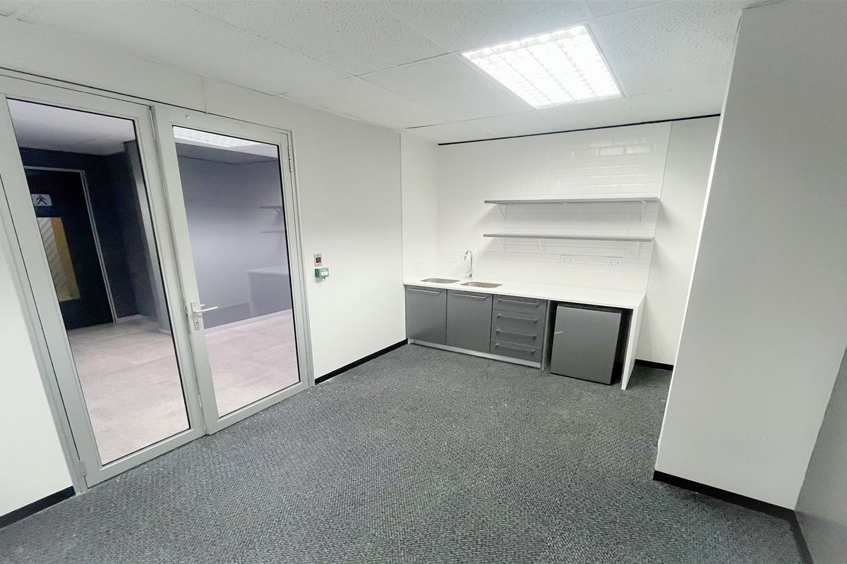 A clean, modern office space featuring a compact kitchenette with built-in cabinetry, sink, and floating shelves, finished with neutral tones and carpet flooring.