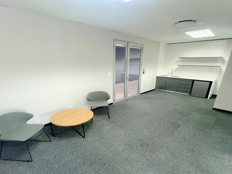 Modern office space featuring a functional kitchenette with built-in cabinetry, sink, and shelving, complemented by neutral finishes and glass double door access.