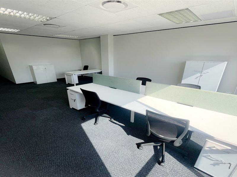 A bright, open-plan office space featuring neutral walls, carpeted flooring, and integrated desk partitions, suitable for a professional work environment.
