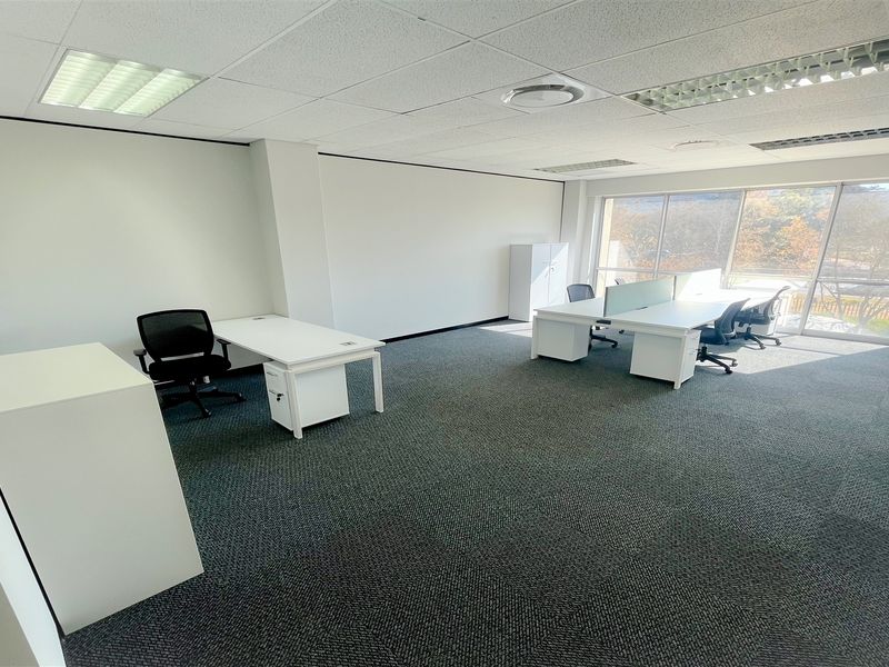 A bright, open-plan office space featuring carpeted flooring, suspended ceilings with recessed lighting, and large windows providing ample natural light.