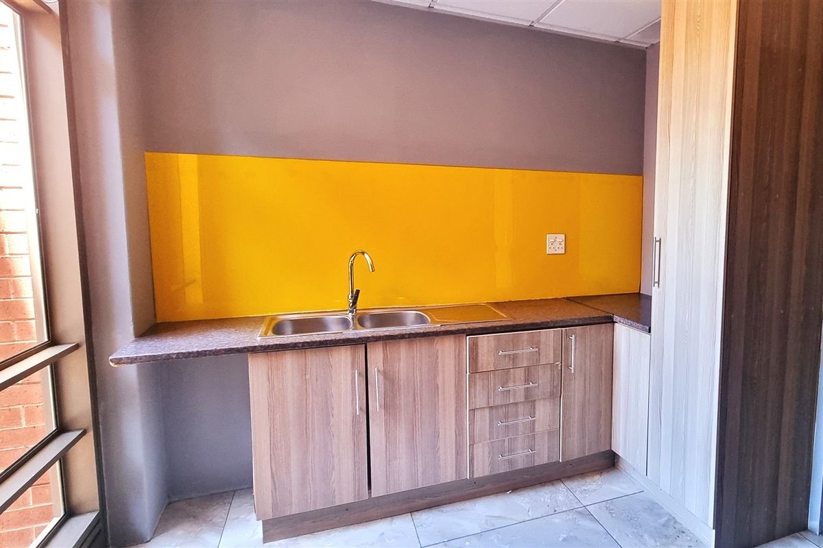 A compact office kitchenette featuring wood-grain cabinetry, a stainless steel double sink, and a vibrant yellow glass splashback.