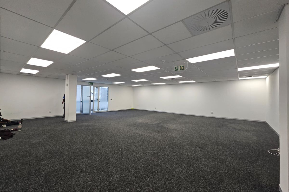 A spacious, open-plan office area featuring a suspended ceiling with recessed lighting, carpeted flooring, and a glass door entrance.