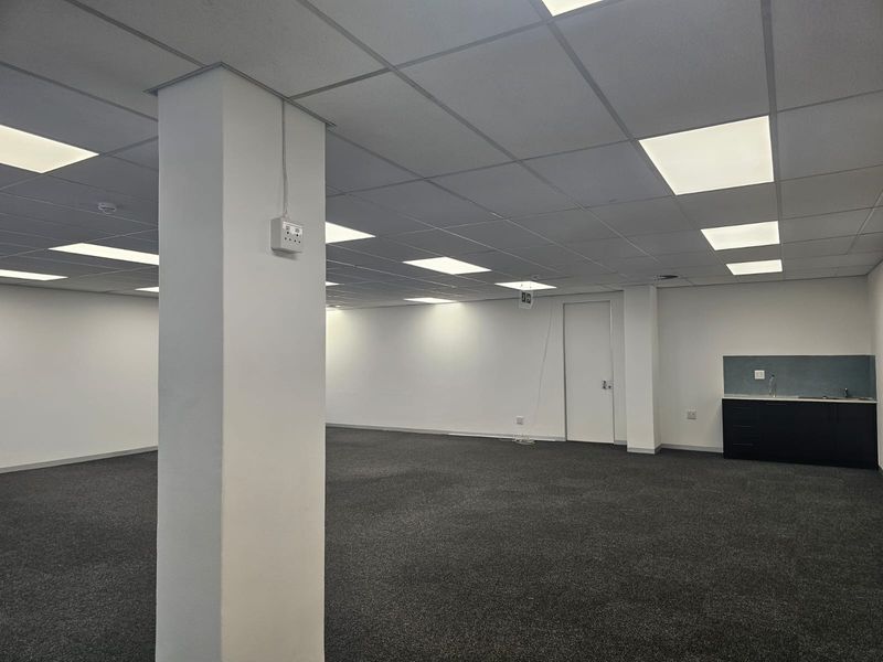 Spacious open-plan office area featuring a suspended ceiling with recessed lighting, dark carpeting, and a compact kitchenette unit.
