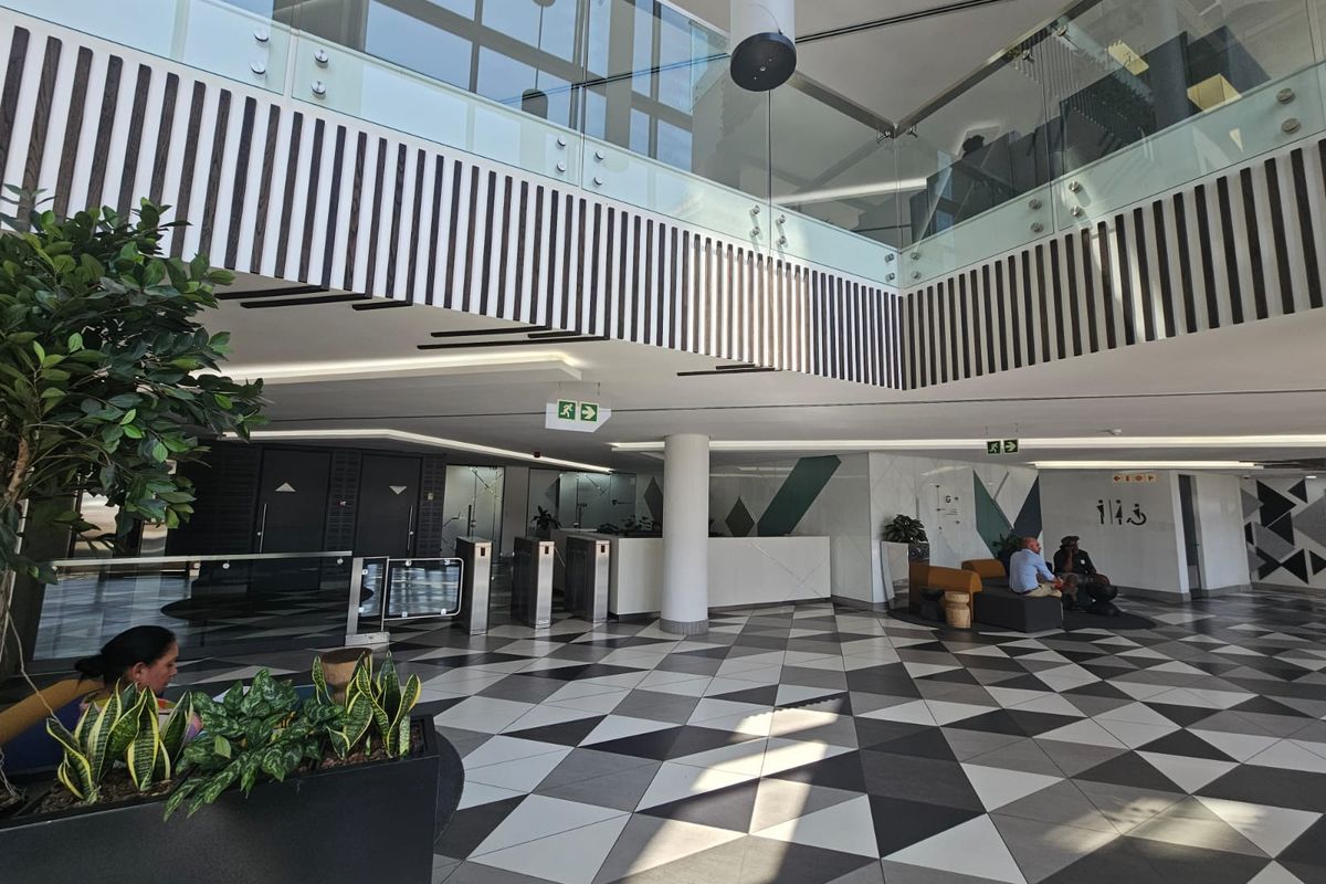Modern office building lobby featuring a double-volume atrium, geometric floor tiling, glass balustrades, and secure turnstile access.