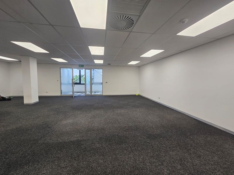 A spacious, empty open-plan office area featuring a suspended ceiling with recessed lighting, air conditioning vents, and dark grey carpeting.