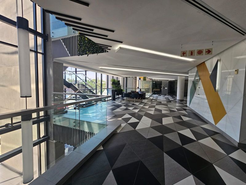 Modern office building hallway featuring geometric floor tiling, glass balustrades, and expansive floor-to-ceiling windows providing ample natural light.
