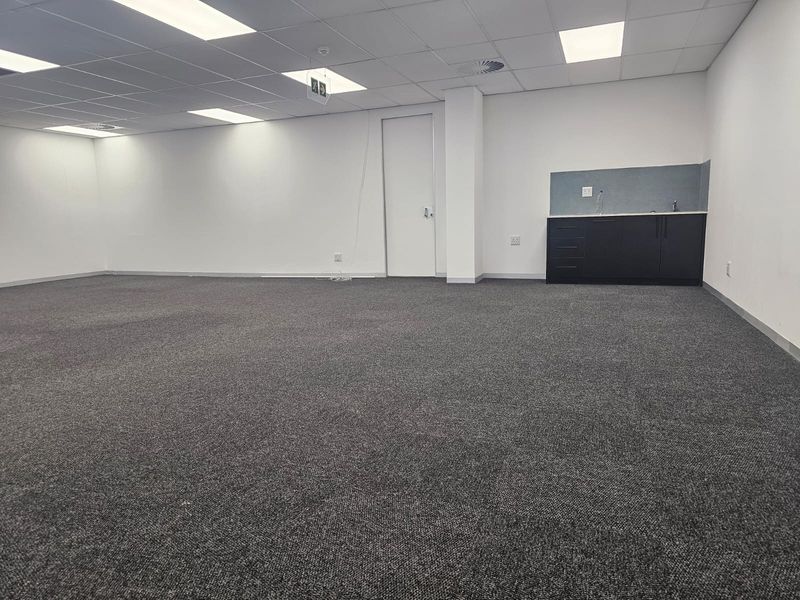 A spacious, open-plan office area featuring neutral carpeting, a suspended ceiling with recessed lighting, and a compact kitchenette unit.