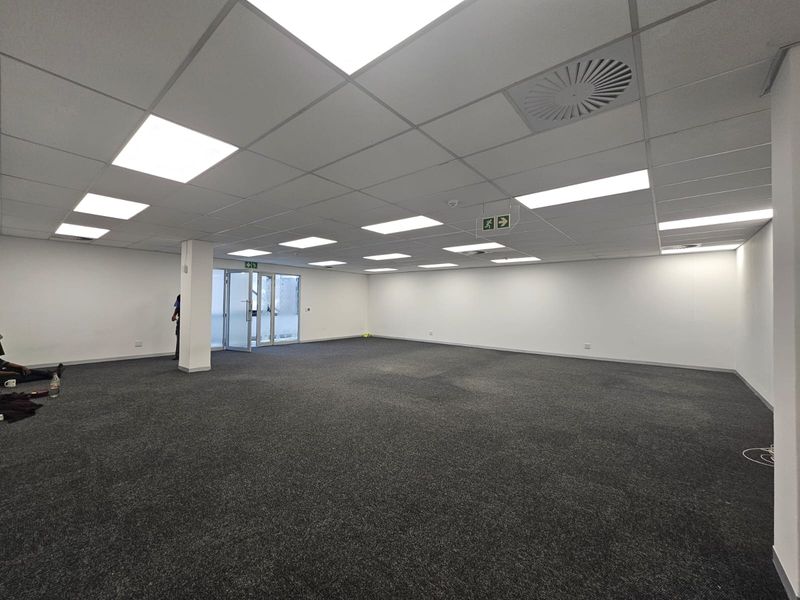 A spacious, open-plan office area featuring a suspended ceiling with recessed lighting, carpeted flooring, and a glass door entrance.