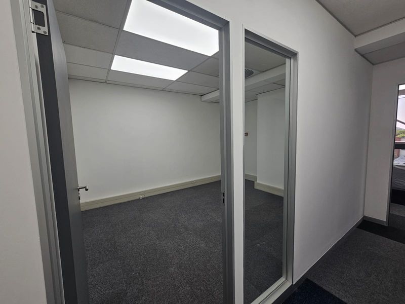 A clean, vacant office space featuring neutral carpet tiles, a suspended ceiling with recessed lighting, and modern aluminium-framed glass partitioning.