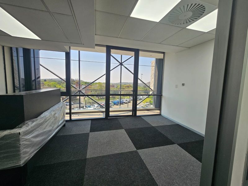 A bright, modern office space featuring carpet tiles, a suspended ceiling with recessed lighting, and large windows offering an exterior view.