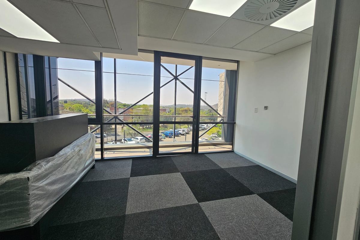 A bright, modern office space featuring carpet tiles, a suspended ceiling with recessed lighting, and large windows offering an exterior view.