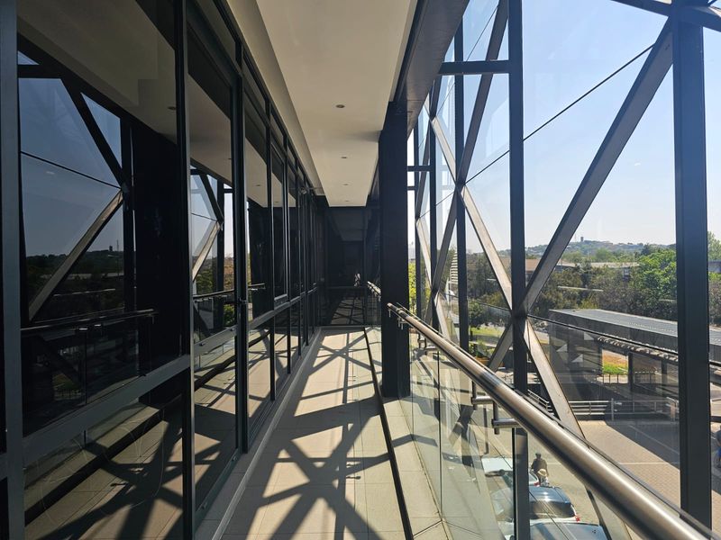 A modern, covered balcony featuring a glass balustrade, metal handrail, and extensive floor-to-ceiling glass windows overlooking the surrounding office park area.