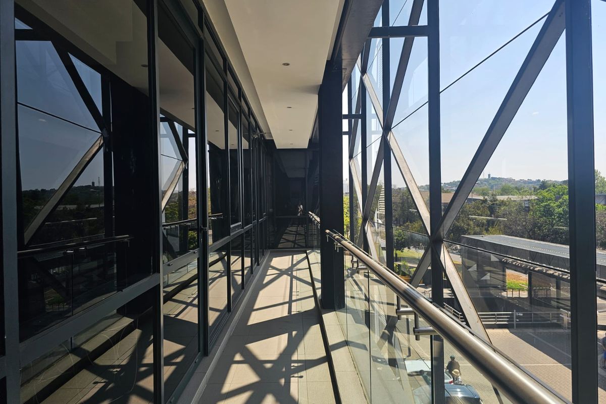 A modern, covered balcony featuring a glass balustrade, metal handrail, and extensive floor-to-ceiling glass windows overlooking the surrounding office park area.