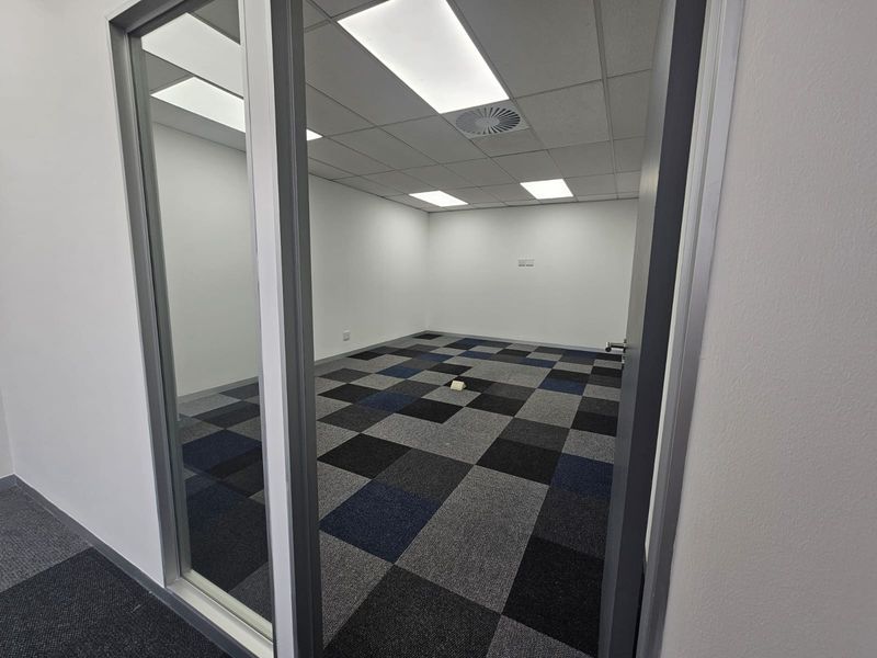 A clean, empty office space featuring modern carpet tiles, a suspended ceiling with recessed lighting, and a glass partition entrance.