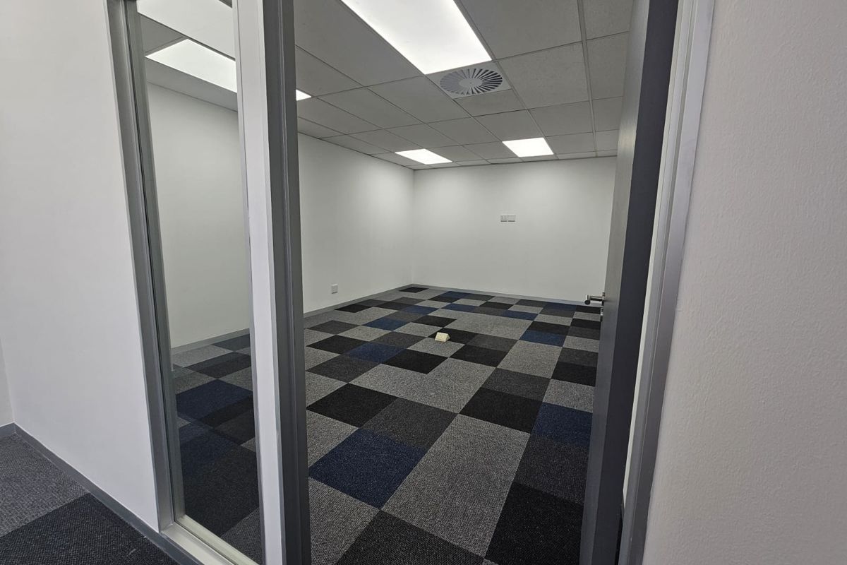 A clean, empty office space featuring modern carpet tiles, a suspended ceiling with recessed lighting, and a glass partition entrance.