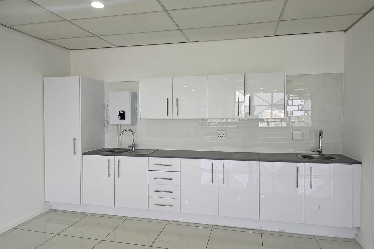 A clean, modern office kitchenette featuring glossy white cabinetry, subway tile backsplash, stainless steel sinks, and tiled flooring.