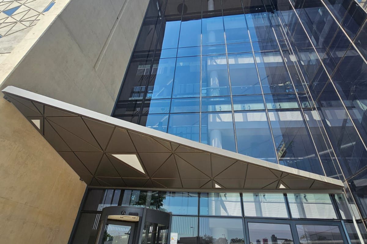Modern office building entrance featuring a prominent glass curtain wall, concrete structural elements, and a geometric metal canopy over the revolving door.
